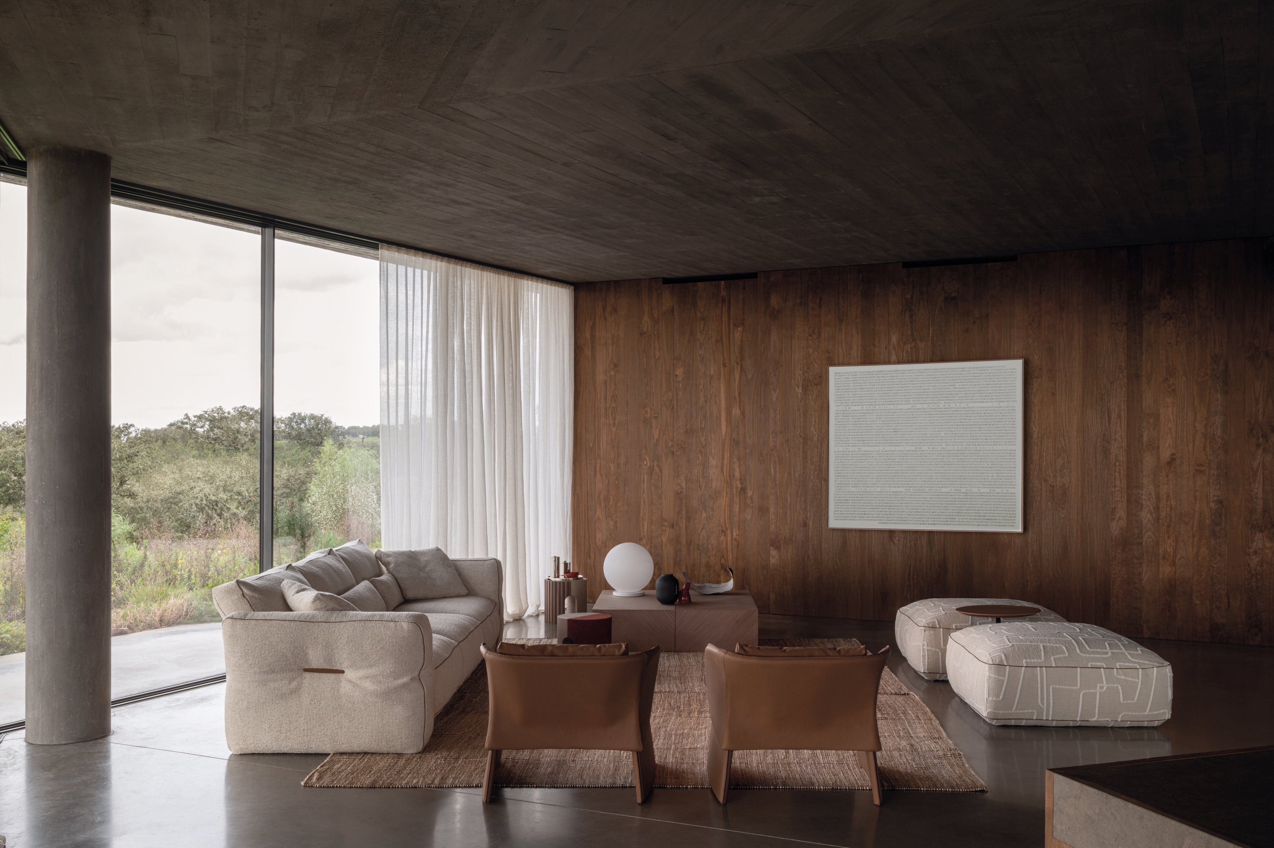 Ambiente living con divano Loden chiaro, due pouf decorativi e sedute in pelle, con grandi vetrate e pareti in legno