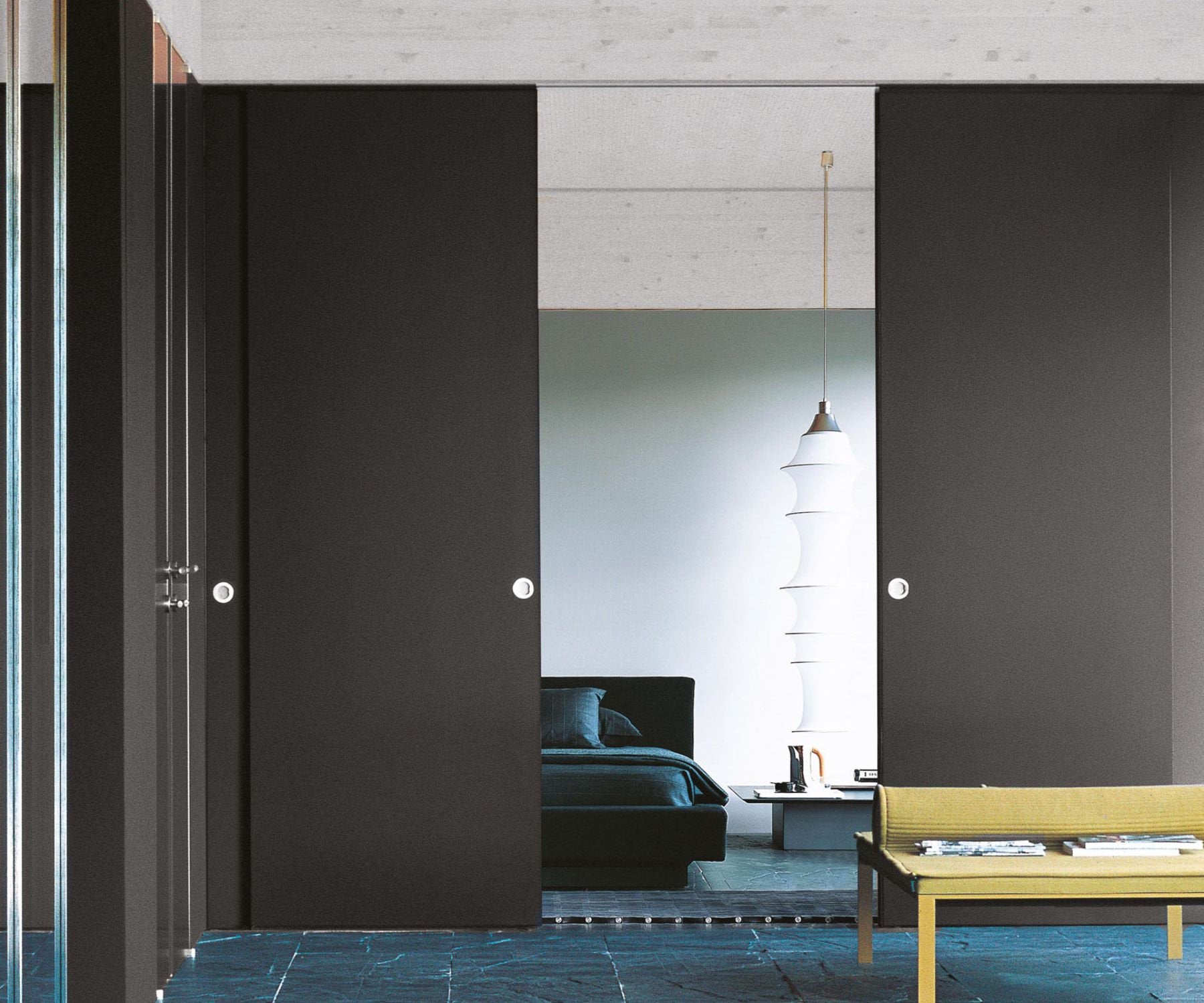 Interno moderno con porte scorrevoli a tutta altezza in finitura nera, maniglie circolari integrate e vista su zona notte con letto basso e lampada a sospensione