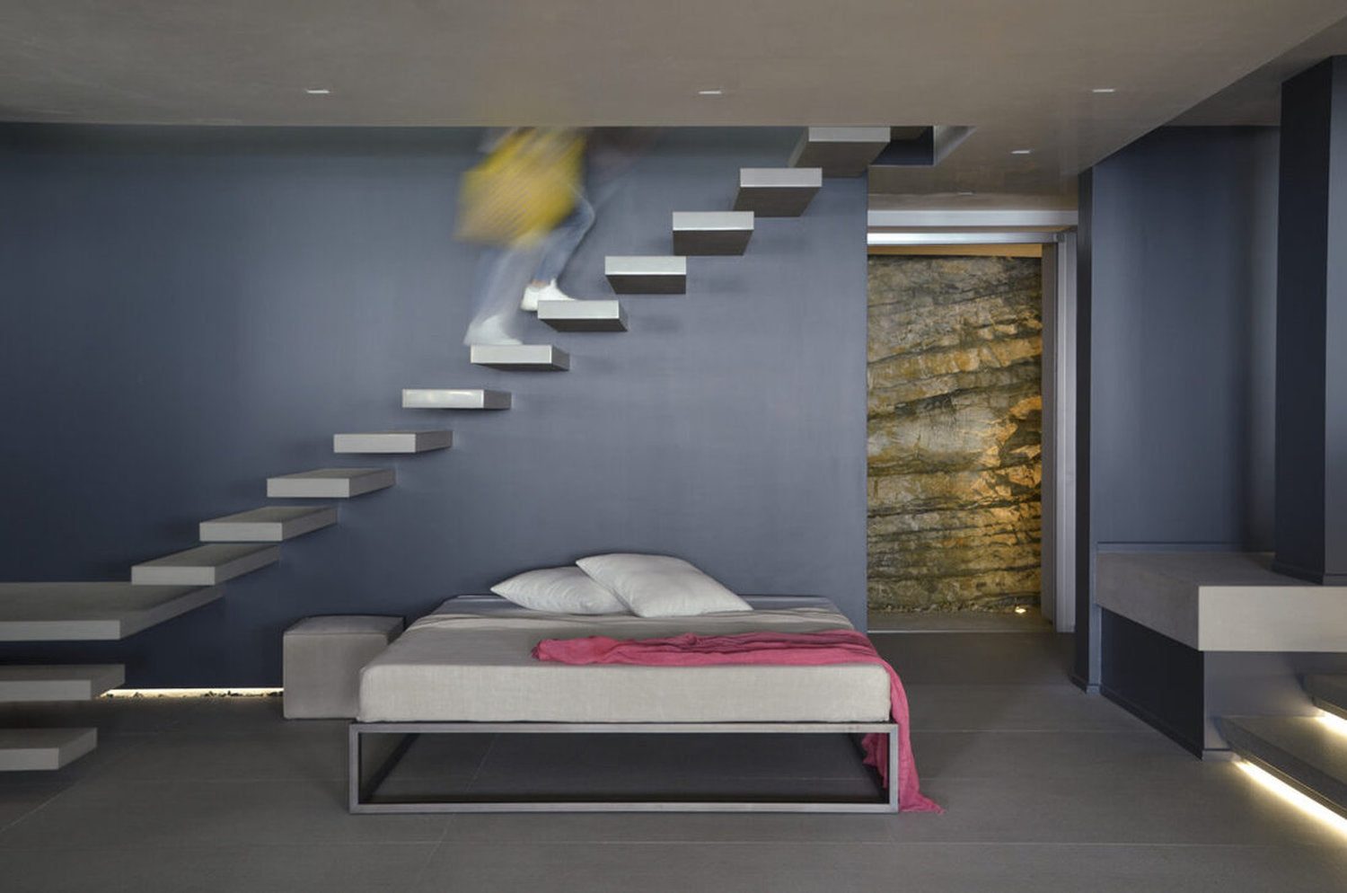 Letto Asha Basic 3.0 sotto scala sospesa in interni contemporanei con parete blu opaco.