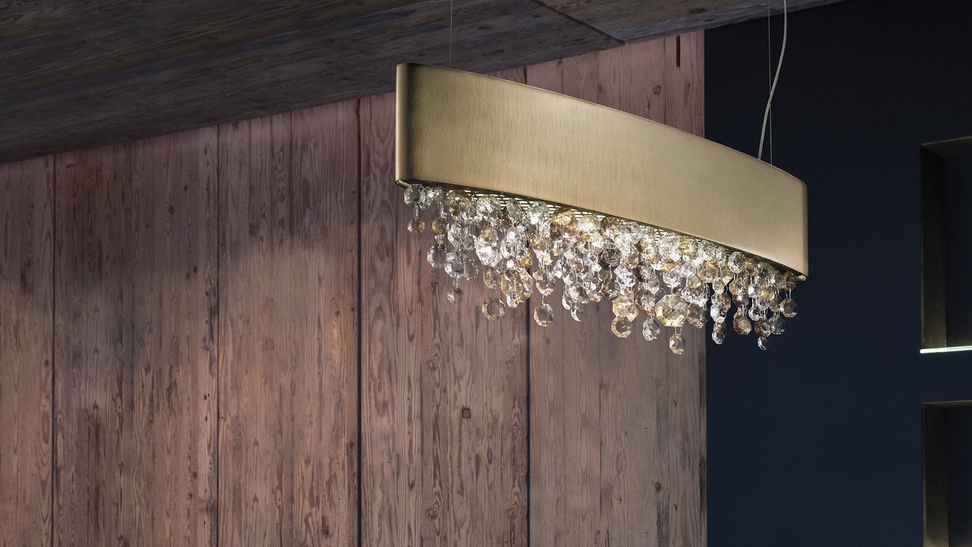 Lampada a sospensione Olà di Masiero con struttura in ottone satinato e pendenti in vetro, installata contro una parete in legno naturale.