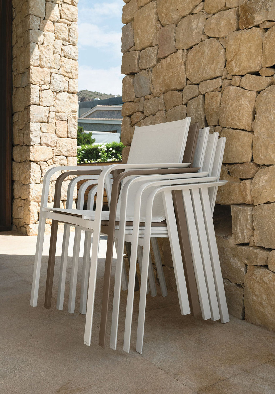 Sedie pranzo outdoor Milo Talenti impilabili in alluminio, in finitura white e dove, accanto a parete in pietra