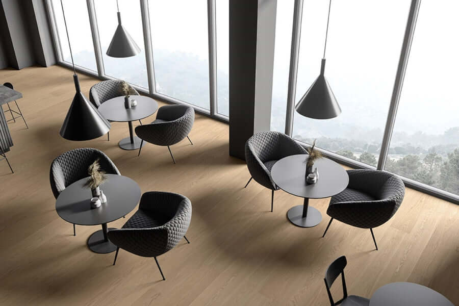 Area contract con tavoli rotondi e poltroncine su pavimento SPC effetto rovere chiaro, con vetrata a tutta altezza