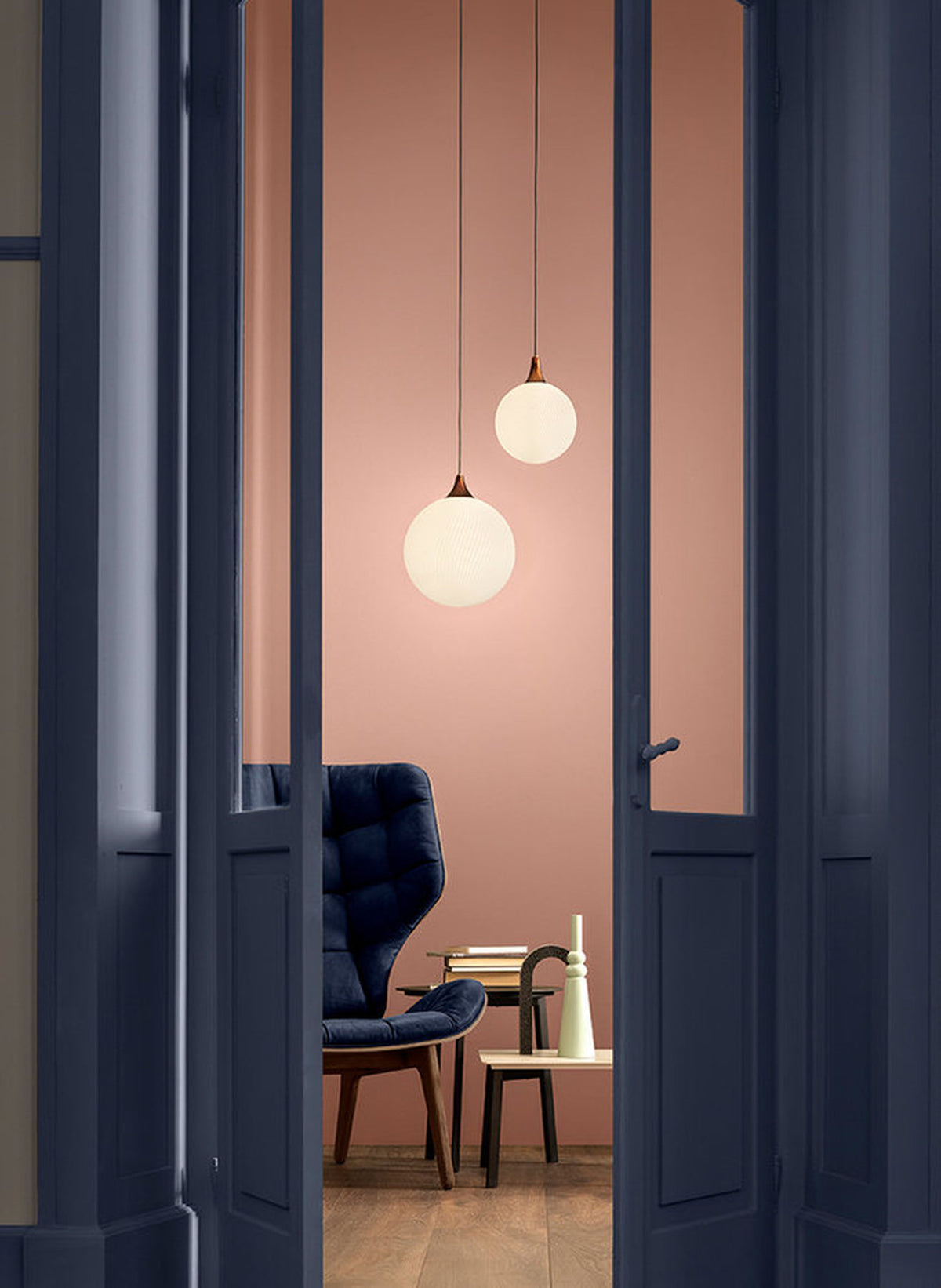 Due lampade a sospensione Tee di Masiero con sfere luminose in vetro bianco, a diverse altezze accanto a una poltrona blu.