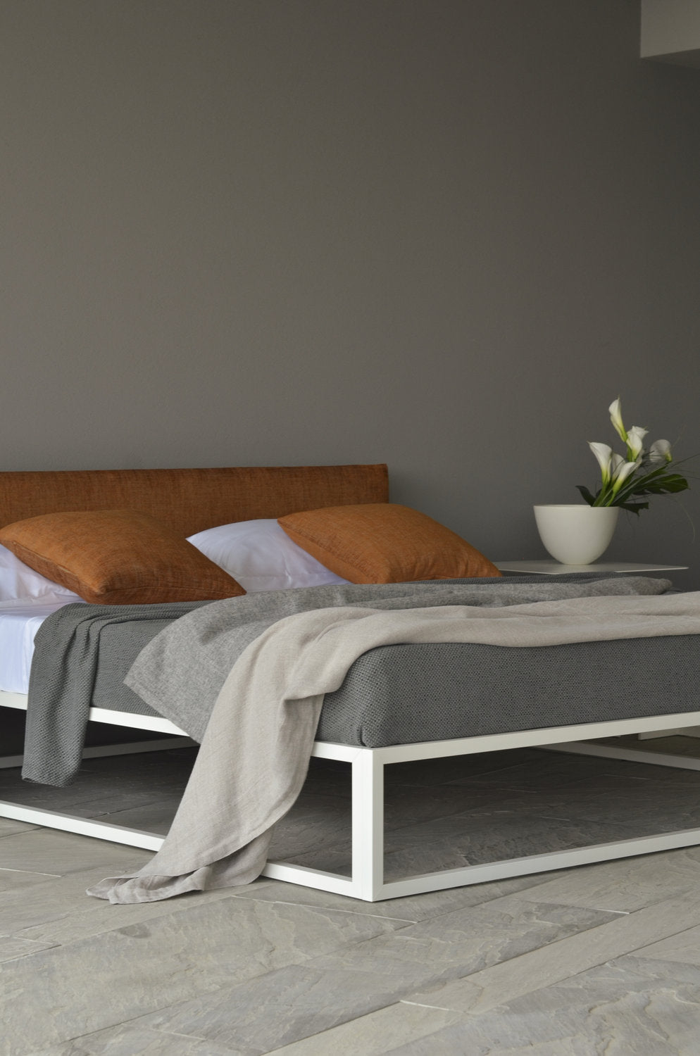 Letto Asha Basic 3.0 con struttura bianca e testata tessile arancio, vista laterale su pavimento in pietra